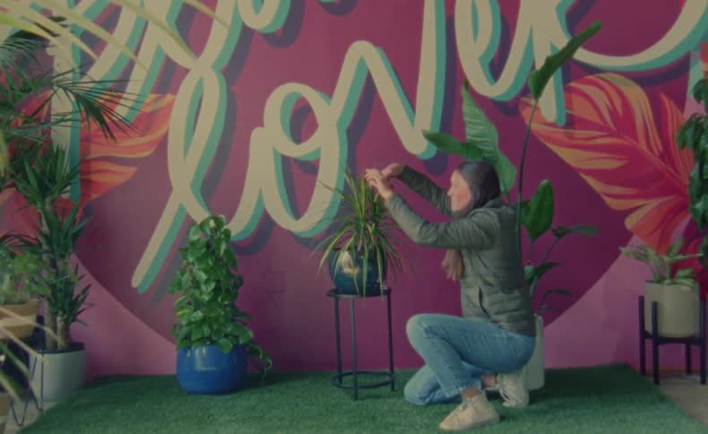 A woman is kneeling down fixing a plant on display with other plants near a painted wall