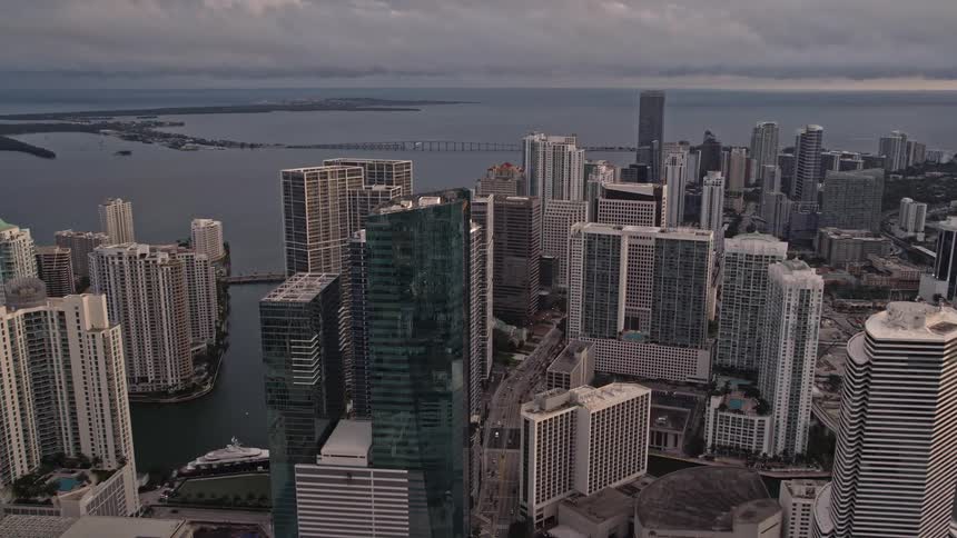 An aerial view at sunset as skyscrapers on the bay shore in Downtown Miami Florida approach and towering buildings are shrouded in cloudy skies