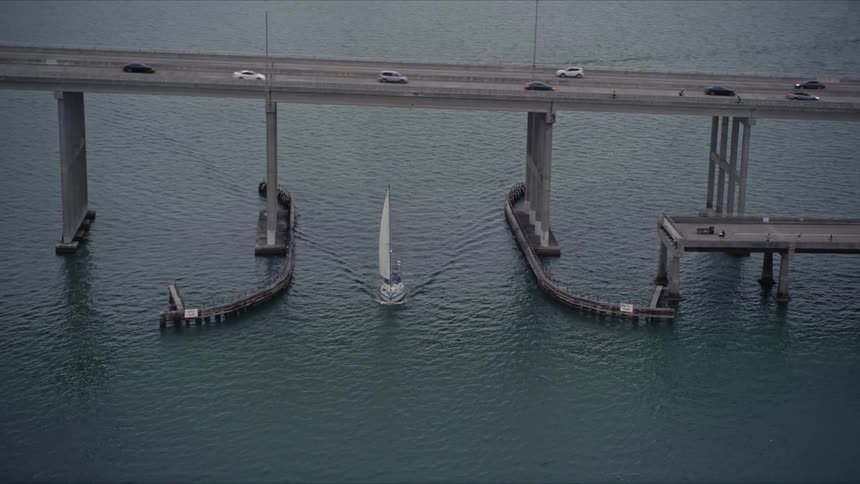 A sailboat passes under a bridge on the Rickenbacker Causeway at sunset in Florida captured in aerial footage