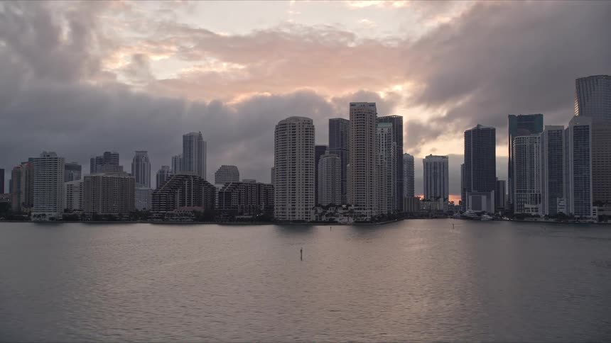 Sunset aerial footage of Miami River approach and Downtown Miami coastal skyline with high-rise towers and skyscrapers against a cloudy sky in Southern Florida