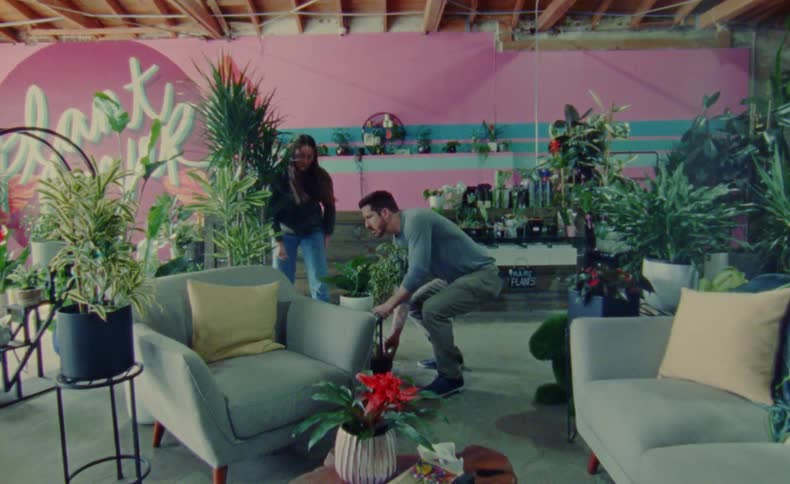 A man and woman work together to carry a large plant into a business