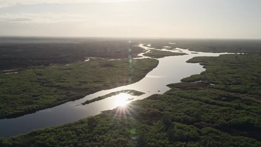 Approaching St Johns River at sunrise in Florida from a 5K aerial view with lush greenery surrounding the river and a beautiful sun flare in the background