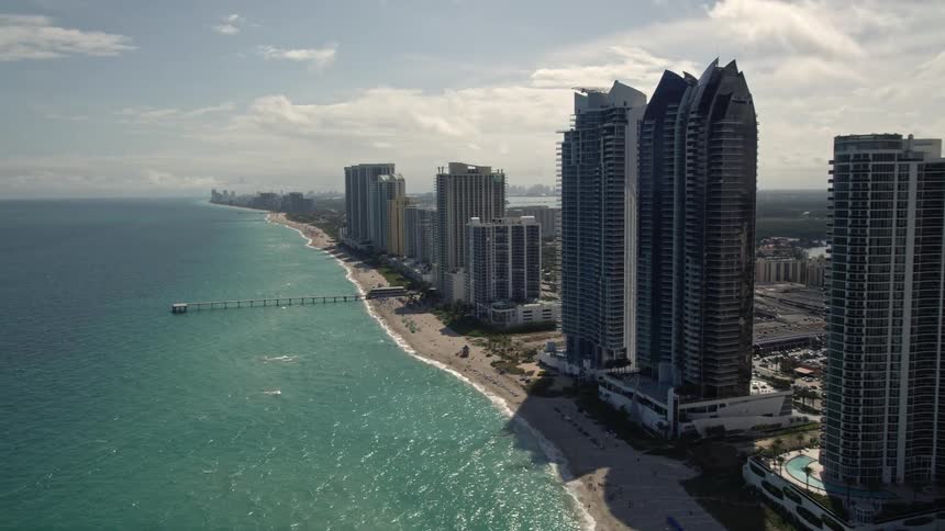Approaching a pier in Sunny Isles Beach Florida fly over the tall beachfront high-rises and coastline residences in an aerial view