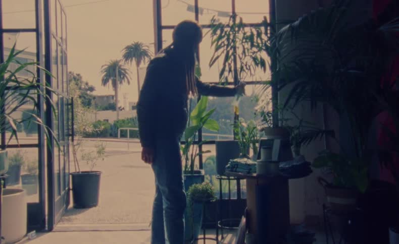 A man is standing near the doorways of a store watering plants with a spray bottle near a table display