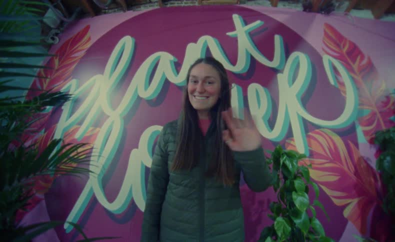 A woman standing in front of a large mural posing for the camera smiling and waving
