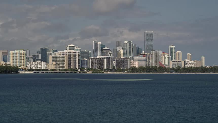 An aerial footage revealing the Downtown Miami skyline from the bay approaching the metropolis city on the shoreline with towers and skyscrapers towering over the coastline on a partly cloudy day in Southern Florida