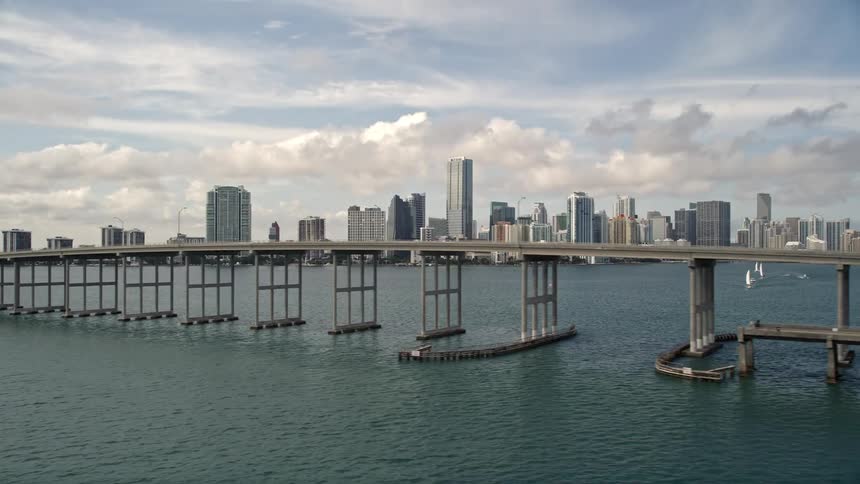Aerial footage of Rickenbacker Causeway showcasing light traffic and Downtown Miami skyline in the background shot during the spring or summer months with partly cloudy skies