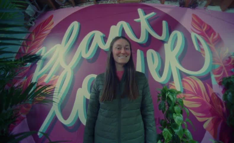 A woman stands in front of her businesss logo while smiling and waving