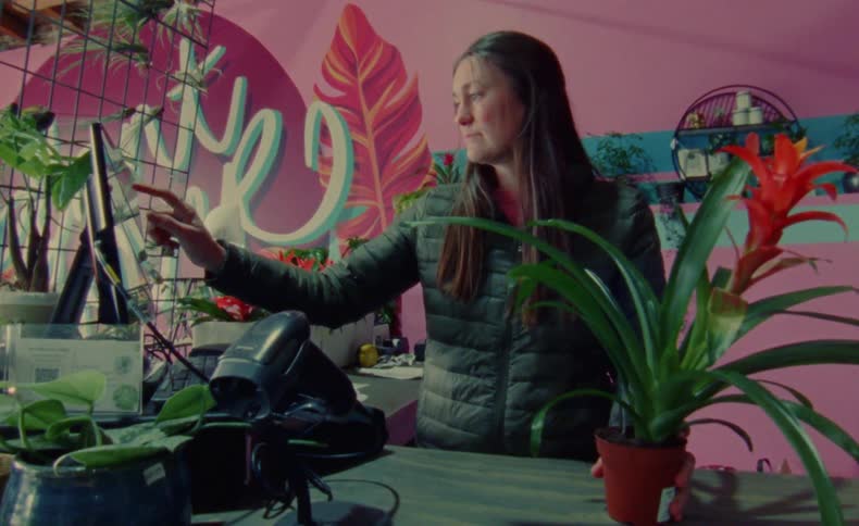 A young lady is in a flower shop using a device to scan a flower and then typing on a desktop computer