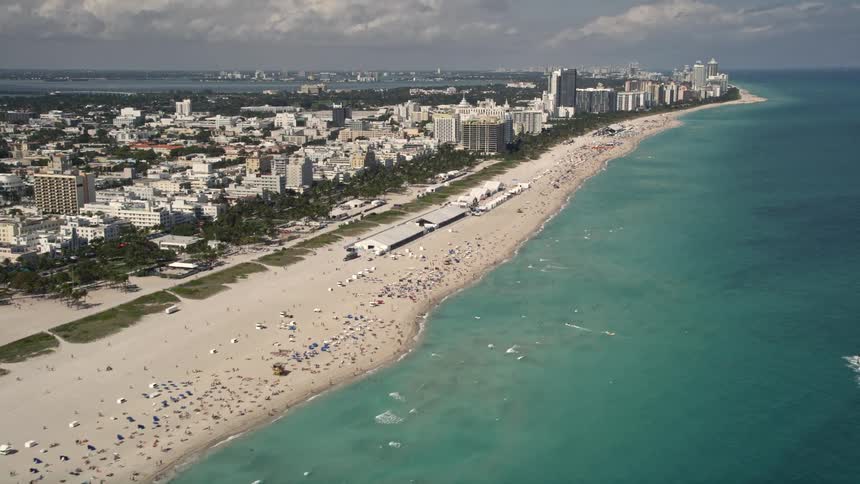 Aerial shot of a lively South Beach Florida tilting up from sunbathers and beachgoers to bustling commercial and residential beachfront buildings against a partly cloudy sky
