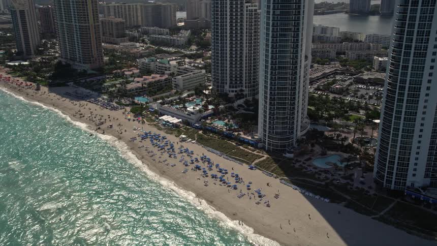 Approaching luxurious beachfront hotel in Sunny Isles Beach Florida with birds-eye-view of sunbathers and umbrellas on the coastline