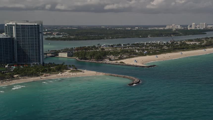 Fly over Bal Harbour inlet revealing oceanside buildings and a bridge with a cloudy Miami skyline in the background