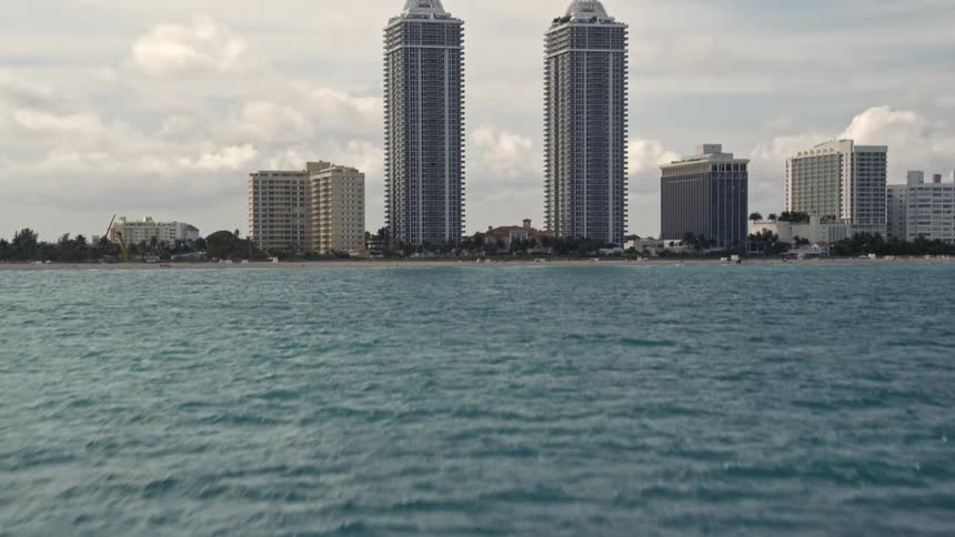 Aerial footage of Miami Beach Florida from a low altitude as the camera tilts from the blue ocean to reveal and approach the stunning Blue and Green Diamonds skyscrapers with partly cloudy skies above