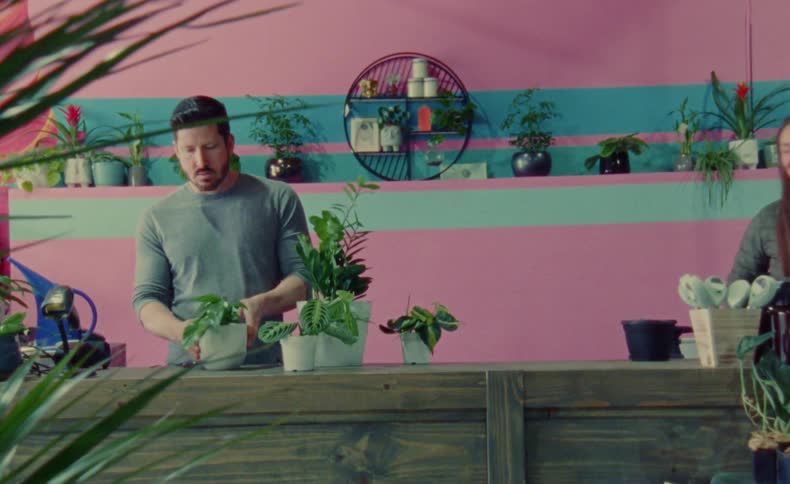 A man has several potted plants in a counter in front of him and adjusts two of them while a woman approaches from the side