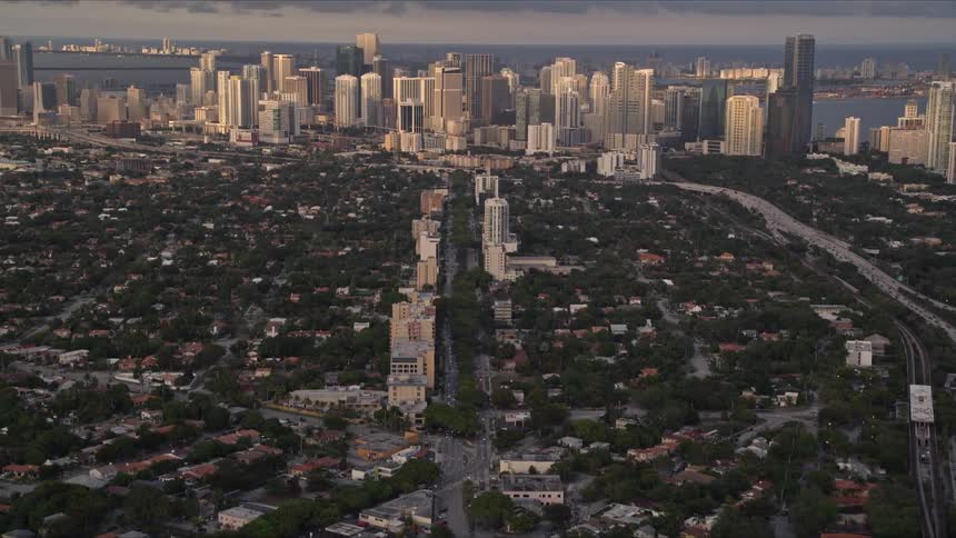 A stunning aerial view of a suburban city gradually revealing the downtown area with towering skyscrapers and a body of water during sunset in Southern Florida