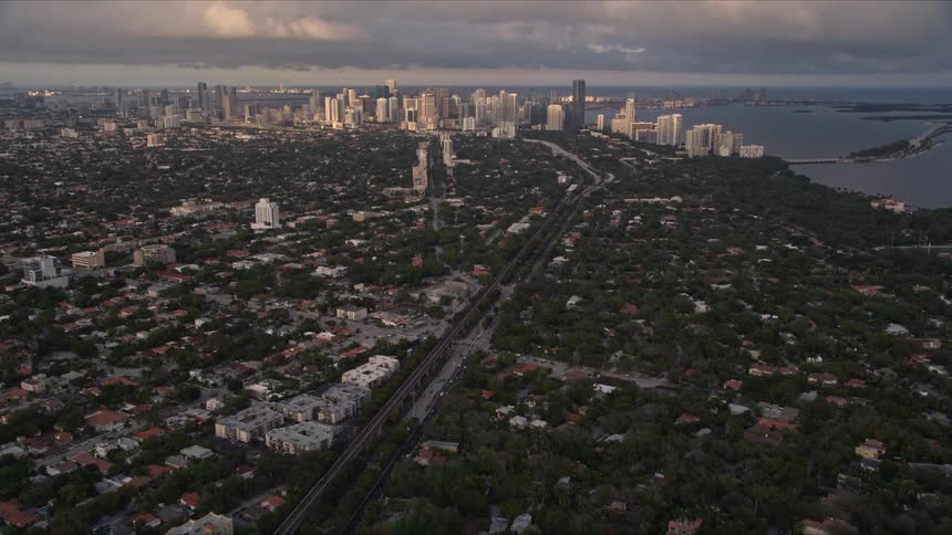 A stunning aerial view of Downtown Miami at dusk as the camera tilts up from the residential Coconut Grove suburbs revealing the impressive city skyline and coastal panorama at sunset