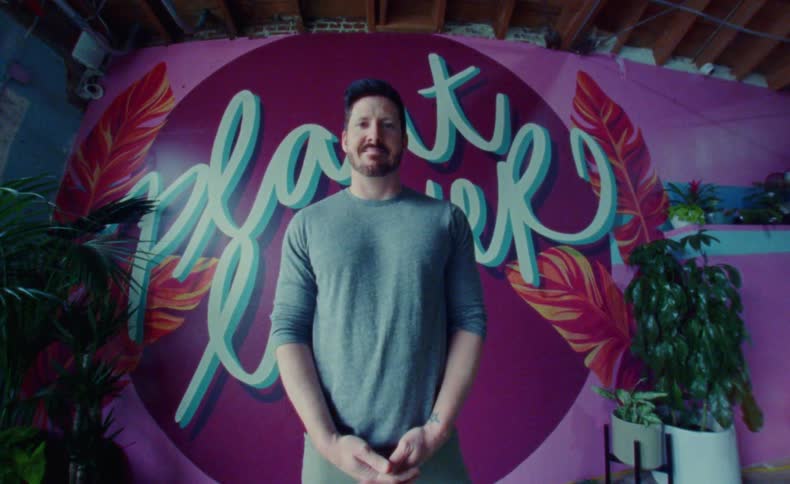 A man stands and poses for the camera in front of his store logo