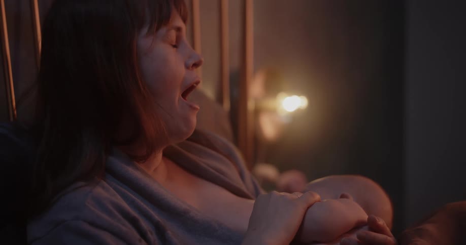 Tired mother yawns while breastfeeding her baby resting in bed on a cozy room at home