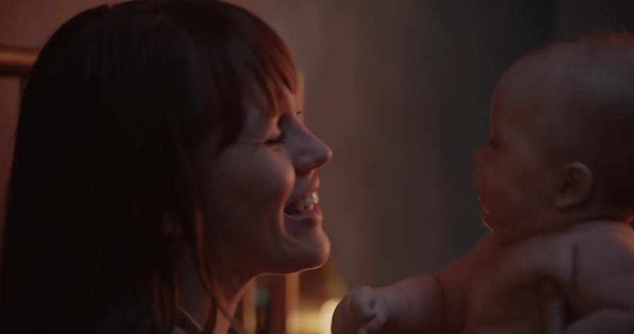 A mother holds up her baby and playfully blows wind in their face while the baby tries to grab her