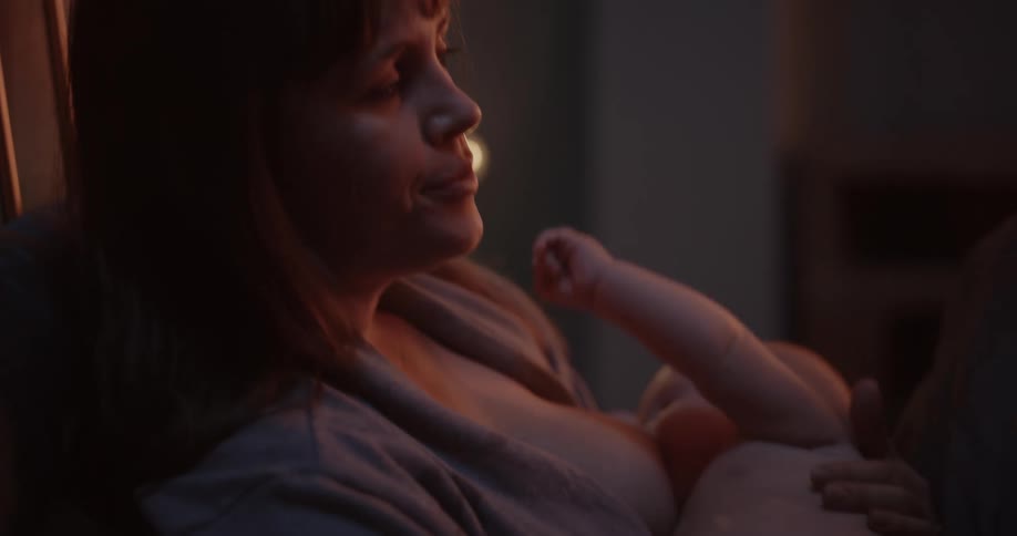 Mother with bangs yawns while breastfeeding baby sitting on bed in the bedroom