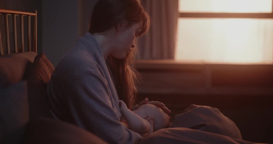 A mother nursing her infant baby in her arms while she sits against pillows on her bed