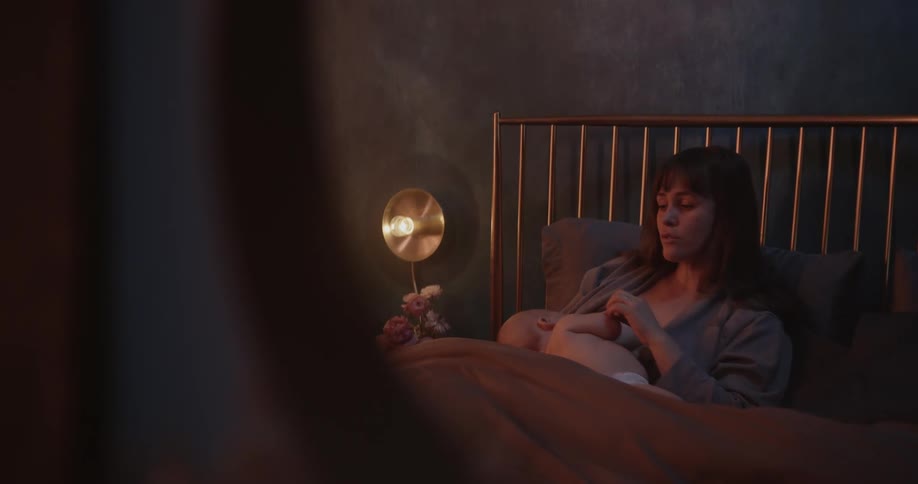 Mother with brown bangs yawns while breastfeeding baby son in the bedroom