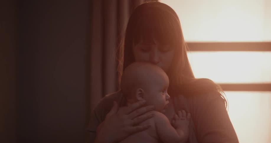 Mother with brown bangs holds newborn baby in her arms while the sun shining through windows