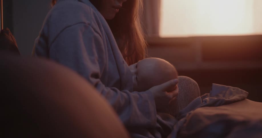 A woman is sitting up in bed with her baby rubbing their head as she nurses the infant