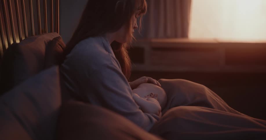 Mother with brown hair breastfeeds newborn baby stroking his head in bedroom