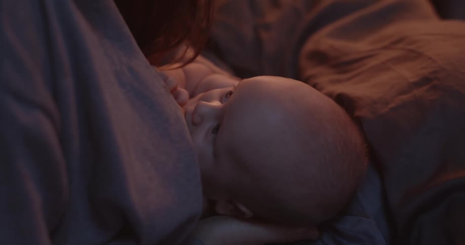 Mother with brown hair breastfeeds newborn baby sitting on bed