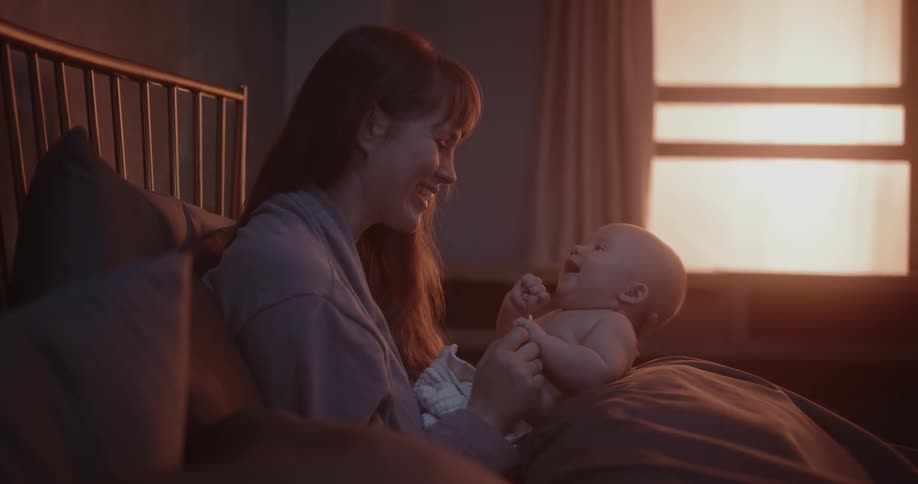 Mother with brown hair holds newborn baby in her arms sitting on bed in bedroom