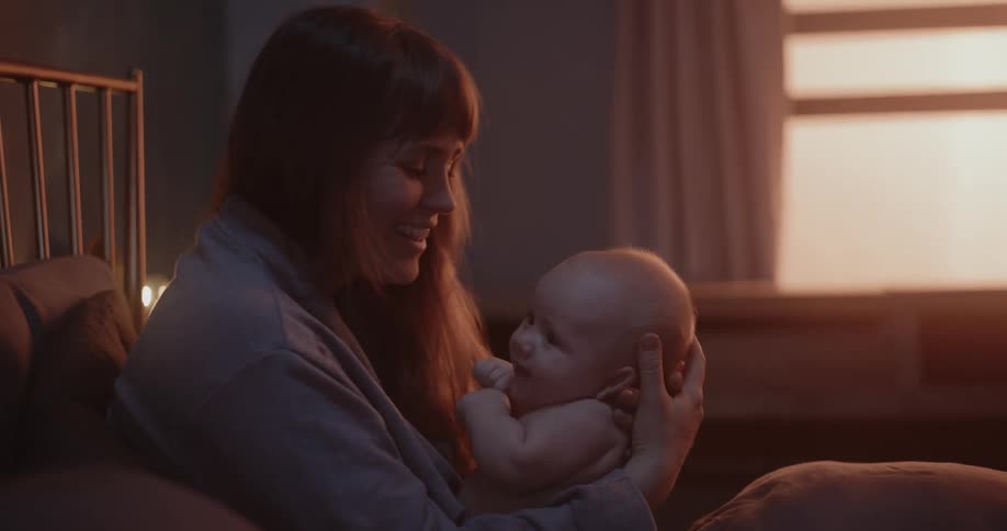 Mother smiles while rocking her baby in bedroom lit by reddish lighting