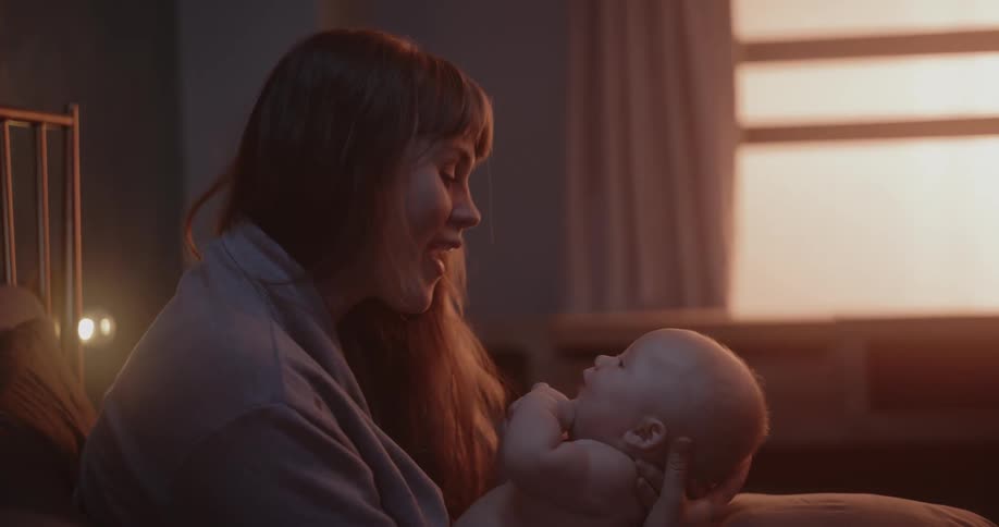 Mother with brown hair holds newborn baby talking and smiling in bedroom
