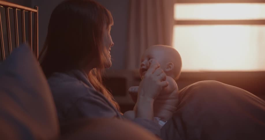 A mother is holding her baby in her lap while they sit in bed and laugh together
