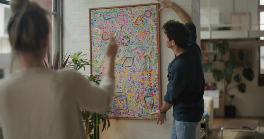 A woman instructs a man as he straightens a painting on a wall before the two high five
