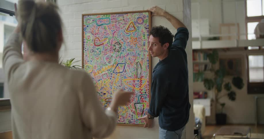 A man playfully tries to streighten an abstract painting on a wall while a woman gives him instructions