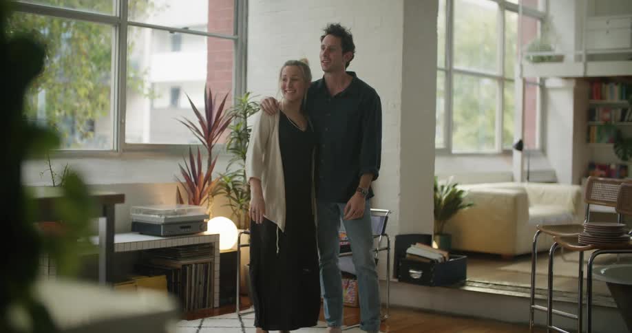 Man in blue shirt and jeans puts down armchair and holds blonde woman with black dress in the living room