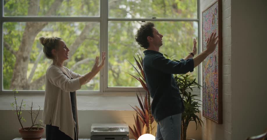 A man is hanging a painting on a wall as a woman stands behind him pointing and giving him instructions and they hug