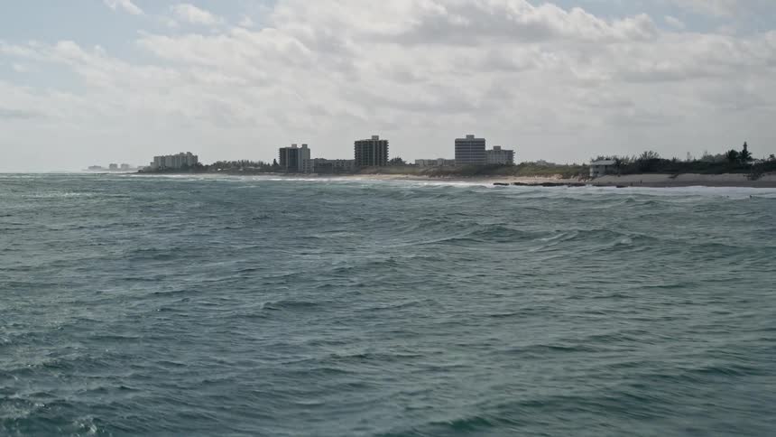 A low flying aerial shot captures ocean waves rolling in to the shore as buildings stand tall along the beachfront in Tequesta Florida
