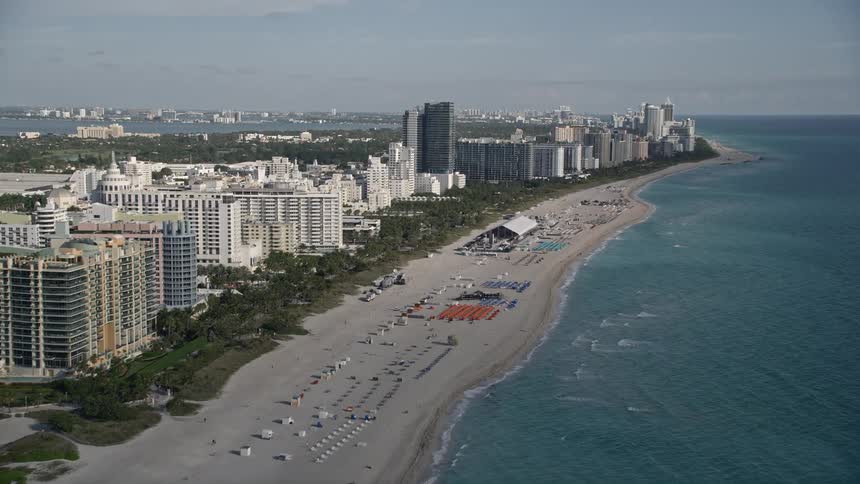 Approaching South Beach hotels aerial footage of Miami Beachs coastline and shoreline with populated beaches and sunbathers enjoying a leisure activity during a partly cloudy spring or summer day