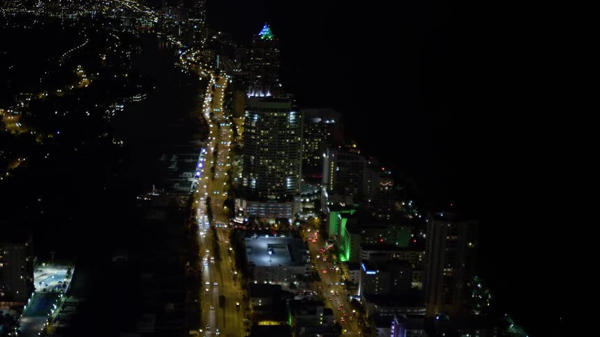 A thrilling night aerial view of Miami Beach Florida as the camera flies over the city lights and busy streets tilting up to approach a luxurious resort hotel