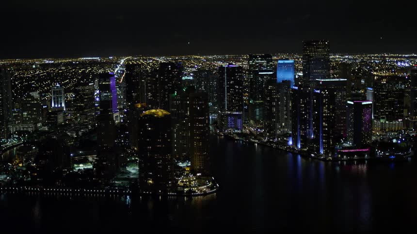 From a side view an aerial footage showcases the clear illuminated nighttime view of tall skyscrapers high-rises and towers in the downtown area of Miami Florida including the shoreline on Brickell Key and the nearby river