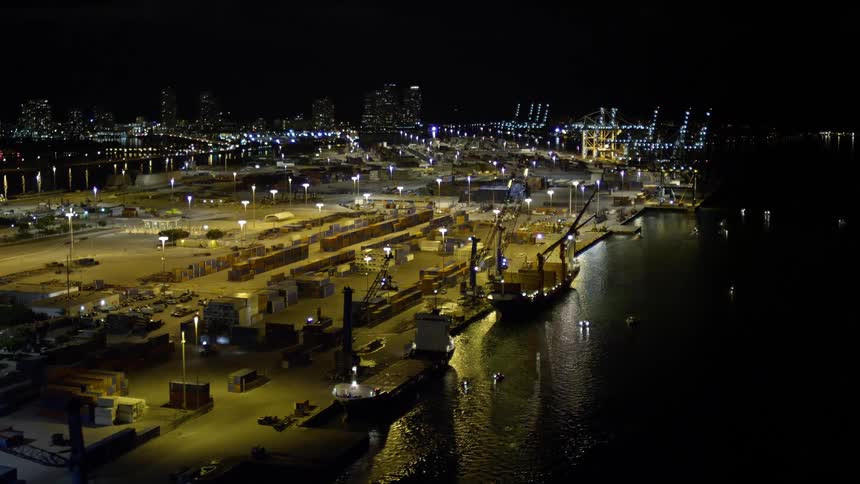 Aerial footage of cargo ships docked below cranes at the Port of Miami in Florida at night with a clear sky and the area lit up by city lights in the distance
