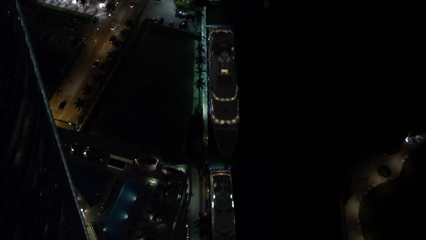 An aerial birds eye orbit of a yacht on the Miami River at nighttime in Downtown Miami showing clear skies and city lights reflecting on the water below