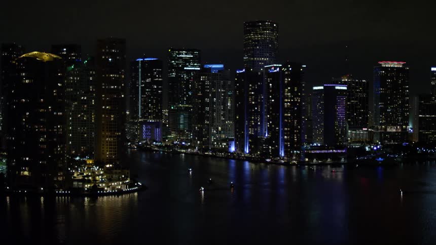 A stunning aerial shot at night over the Downtown Miami Florida captures the tilting-up reveal of countless tall buildings lining the shore of a coastal city