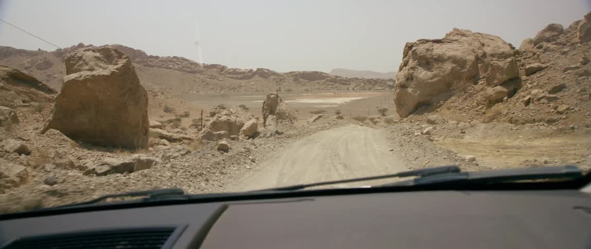 An automobile is traveling on a dirt road through the desert mountains with boulders on the roadside