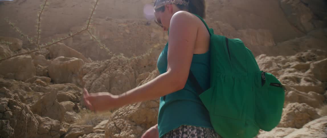 Two women hiking up a rocky mountain with backpacks on and looking at plants.