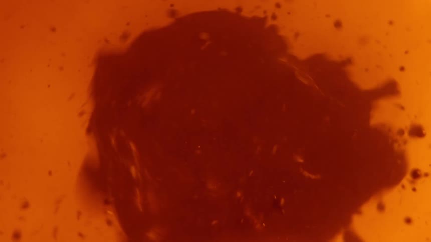 A thick red substance spins and splatters as droplets fly as it moves in an orange liquid atmosphere