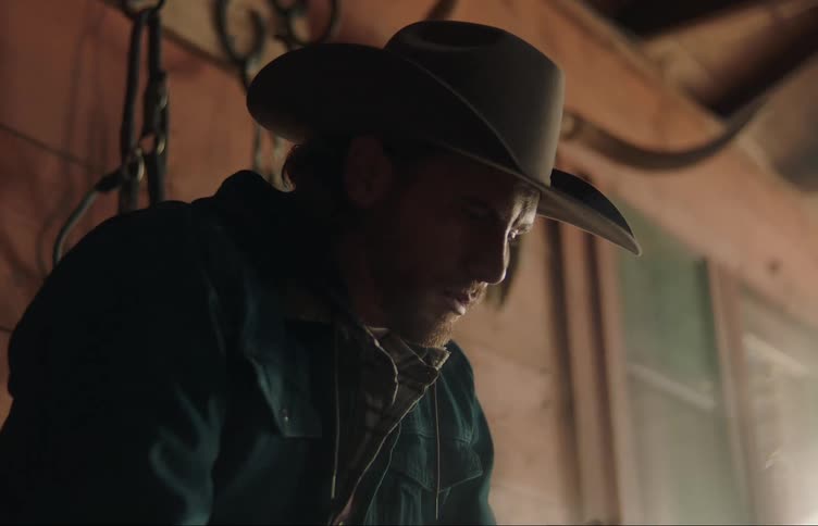 A man is sitting down on a porch with a horned animal skull on the wall as he puts on his cowboy hat and gets up and walks away