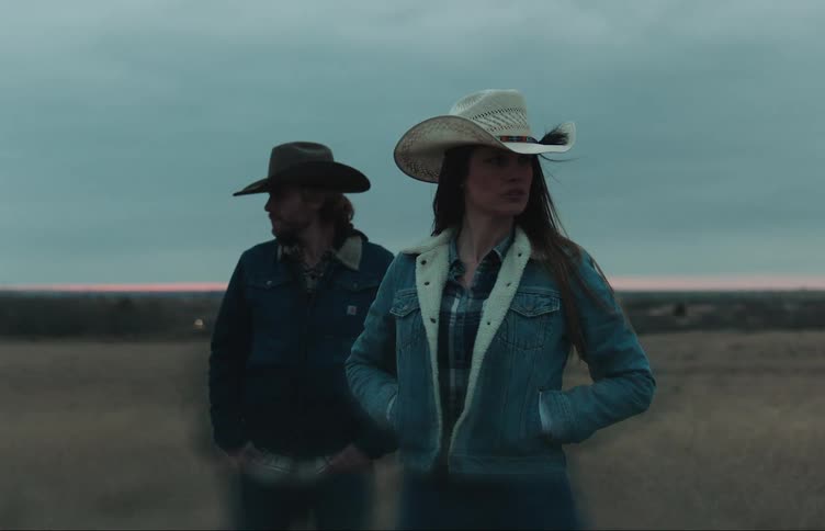 A woman and a man wearing cowboy hats are standing in a rural field with their hands in their pockets as the wind blows their hair